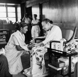 A young woman in a sari kneels and holds the hands of an older woman seated in a chair