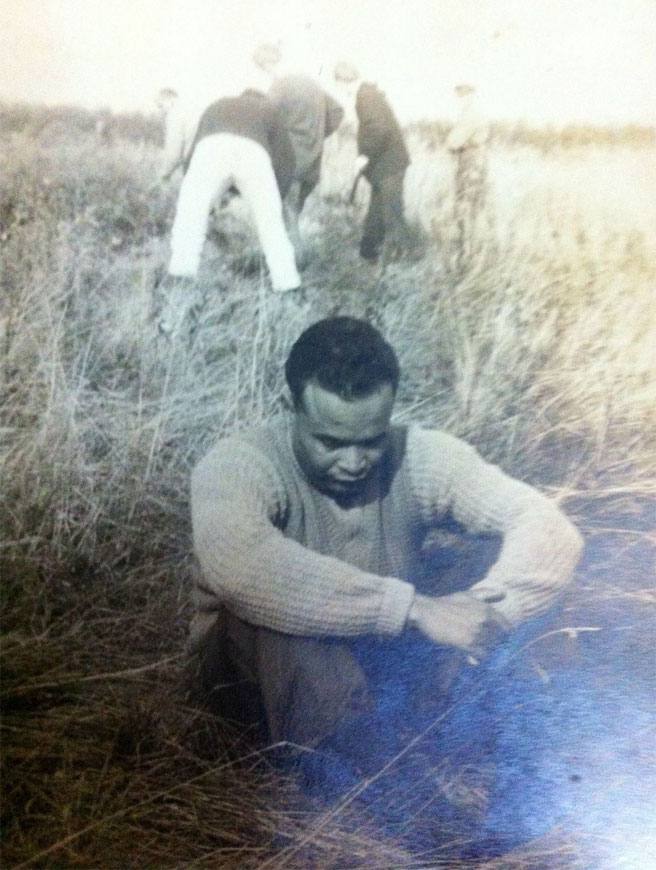 A man in a sweater sitting in tall grass, with others in the field behind him